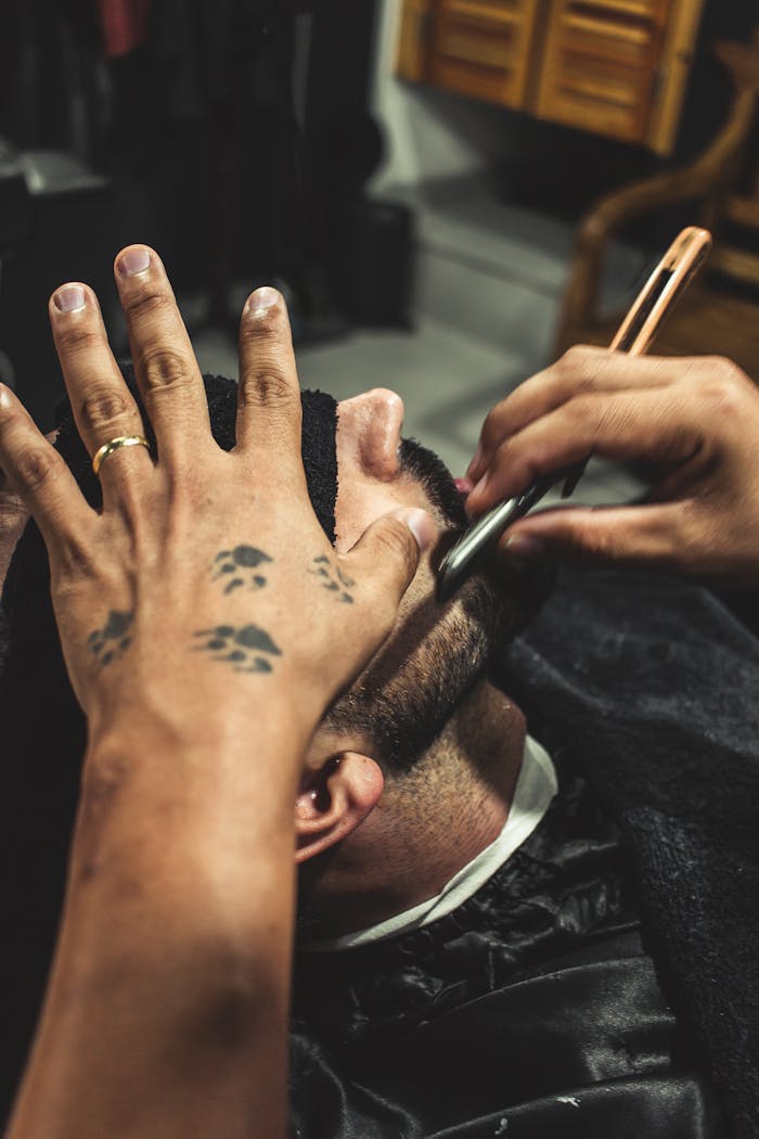 A barber using a straight razor to shave a mans face indoors, showcasing tattooed hands.